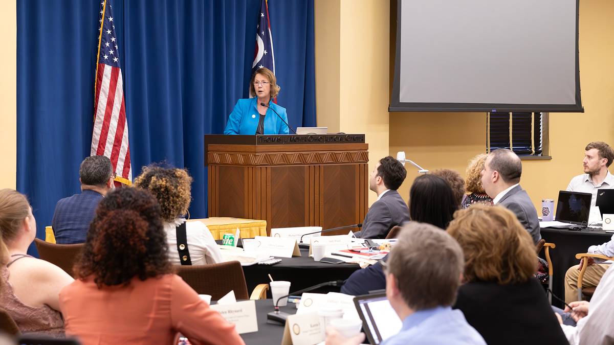 Image of a woman wearing a blue jacket standing at a wooden podium speaking to a roomful of men and women.